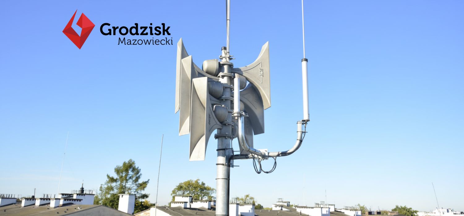 Syreny powiadamiania alarmowego zamontowane na dachu budynku, w tle widoczne dachy innych budynków oraz niebieskie niebo. W lewym górnym rogu widoczny logotyp Gminy Grodziska Mazowiecki